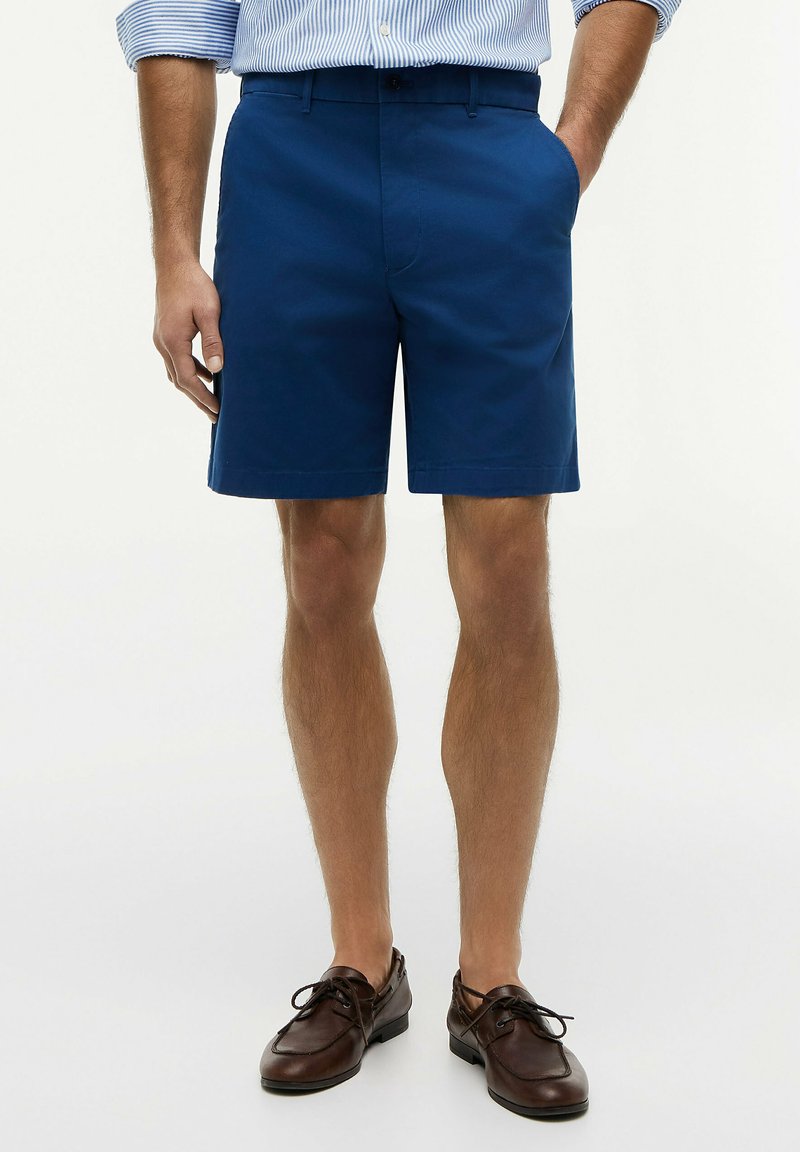Man wearing blue tailored shorts, light blue striped shirt with rolled sleeves, and dark brown leather lace-up shoes.