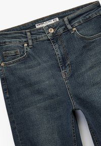 Mörkblå denimjeans med en framficka, kopparknappdetalj och kontrasterande sömmar. Lappen visar storlek W27, stil "Victoria Crop."