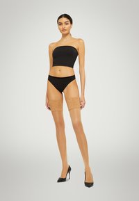 Collant beige trasparente alti fino alla coscia con bordino in pizzo, indossati con un top bandeau nero e biancheria intima nera, abbinati a tacchi alti neri.