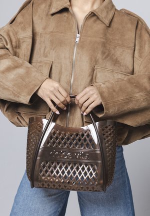 Person som har på seg en brun semsket jakke og blå jeans, og holder en gjennomsiktig brun plastveske med rutemønster og håndtak.