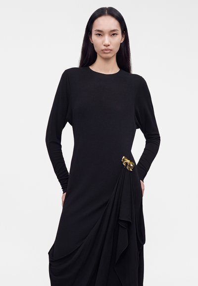 Femme aux longs cheveux noirs porte une robe noire à manches longues avec un détail drapé et un petit nœud doré sur le côté inférieur.