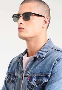 Ochelari de soare negri cu lentile verzi, detalii din metal, formă rectangulară; purtați cu o jachetă din denim peste o cămașă roz deschis.