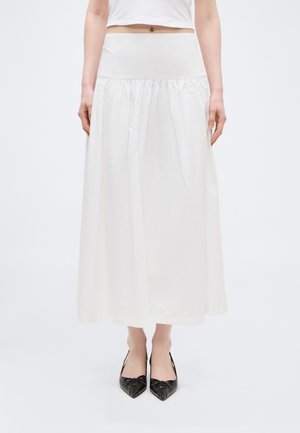 Žena oblečená v bielej midi sukni s vysokým pásom a nariasením, kombinovanej s čiernymi lodičkami s ostrou špičkou, na jednoduchom pozadí.