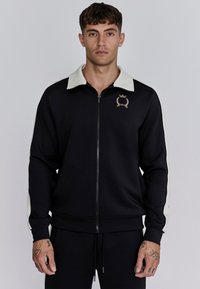 Veste noire zippée avec col crème et accents latéraux, arborant un logo doré sur la poitrine. Fabriquée à partir d'un tissu lisse et extensible.