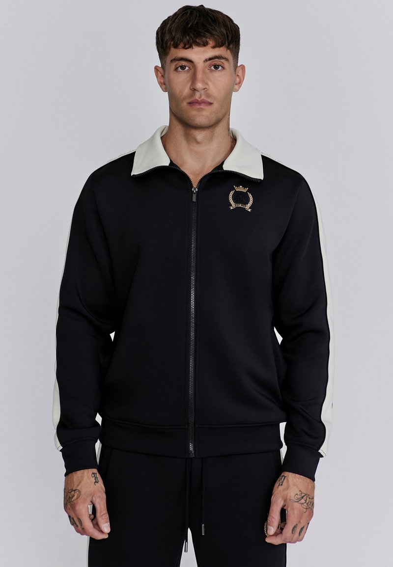 Veste noire zippée avec col crème et accents latéraux, arborant un logo doré sur la poitrine. Fabriquée à partir d'un tissu lisse et extensible.