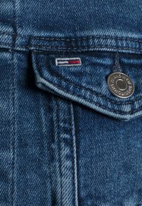 Denim tyg i mörkblått med en ljus tvätteffekt, med en silverknapp och broderad logotyp med röda och vita detaljer.