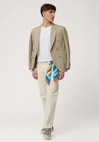 Blazer beige de doble botonadura sobre una camiseta blanca, pantalones crema y una bufanda de colores con un patrón atada a la cintura, combinada con zapatillas.