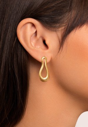 Goldene Tropfohrringe mit einem wellenförmigen Design, das eine glatte Textur und ein poliertes Finish aufweist, hängen in der Nähe eines gestylten Ohrs.