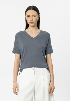 Frau mit glatten dunklen Haaren, die ein kurzärmeliges graues V-Ausschnitt-Shirt trägt, das in hoch taillierte weiße Hosen gesteckt ist, steht vor einem schlichten Hintergrund.