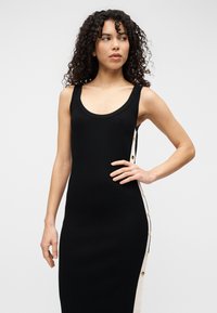 Μαύρο πλεκτό αμάνικο φόρεμα με εφαρμοστή σιλουέτα, διακοσμημένο με αντίθετη μπεζ πλαϊνή λεπτομέρεια και χρυσά κουμπιά.