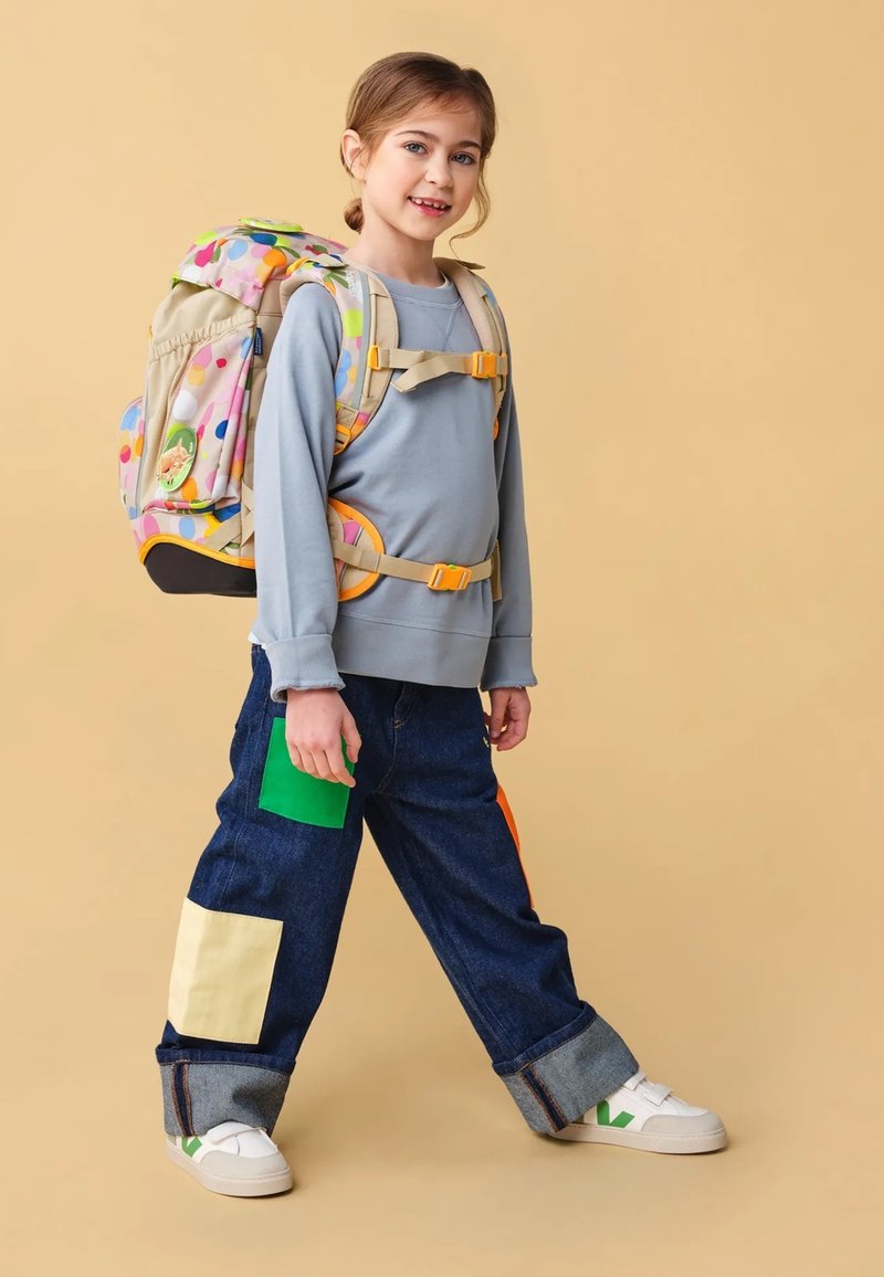 Zaino multicolore con pattern a cerchi, base beige, spallacci regolabili; felpa grigia, jeans blu con grandi toppe colorate, sneakers bianche.