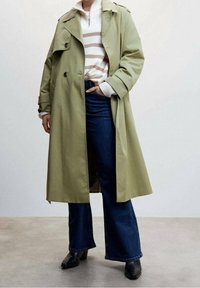 Olijfgroene trenchcoat met dubbele rij knopen, brede revers en schouderepauletten, gedragen over een gestreepte trui en uitlopende spijkerbroek.