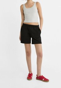 Osoba v světle šedém bezramenném topu, černých šortkách s elastickým pasem, s rukama v kapsách a v červených teniskách s bílými akcenty.