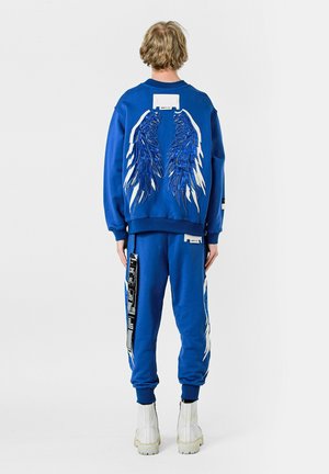 Conjunto de sudadera y pantalones azul, con diseños de alas blancas y azules en la espalda y los lados. Hecho de un tejido suave con puños acanalados.
