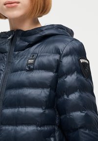 Γυναίκα που φοράει μπλε σκούρο παντελόνι με κουκούλα και μπαλώματα Blauer USA στο στήθος και το μανίκι, με μερική θέα του προσώπου και των μαλλιών.