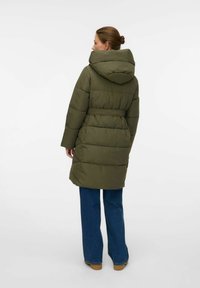 Kvinde iført en olivengrøn belted puffervinterjakke med hætte, blå jeans og brune sko, stående med ryggen til mod en hvid baggrund.