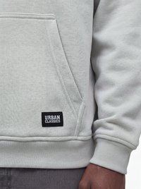 Poche de sweat-shirt gris clair avec étiquette noire "Urban Classics" à l’avant, portée par une personne à la peau foncée et portant un pantalon gris foncé.