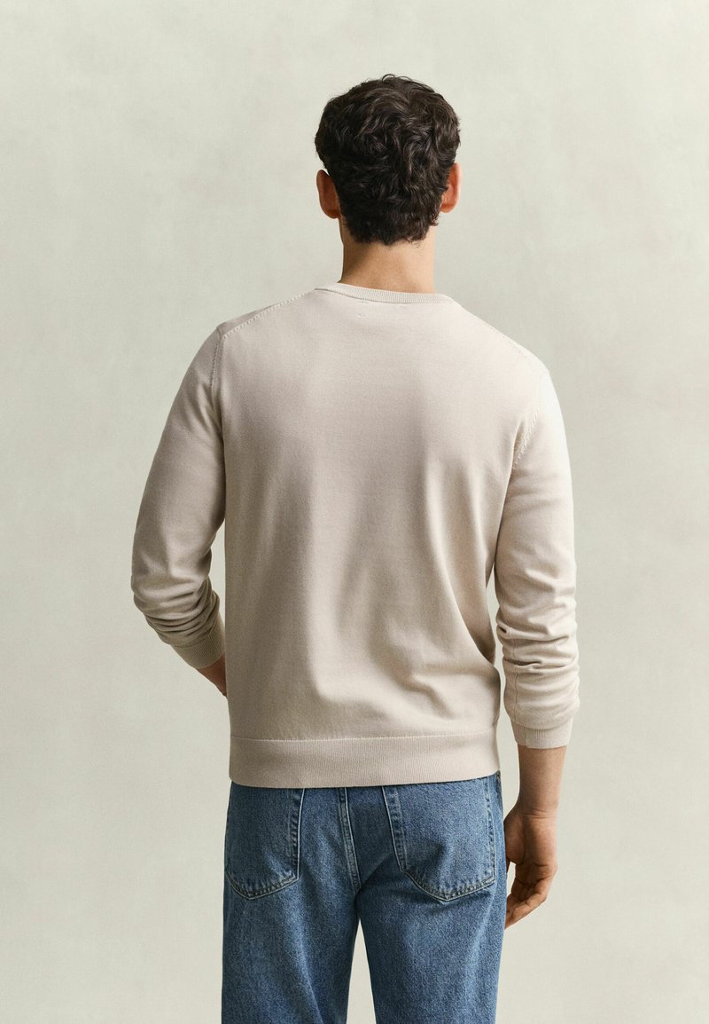 Homme aux cheveux courts, bouclés et foncés, vu de dos, portant une chemise beige clair à manches longues et un jean bleu denim sur un fond uni.