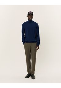 Marineblauer Rollkragenpullover, lockere Passform, kombiniert mit olivgrünen Hosen. Eine schwarze Mütze mit pinkem Text und schwarze Schuhe vervollständigen das Outfit.
