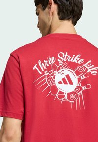 Červené bavlnené tričko s bielou grafikou na chrbte, zobrazuje bowlingovú tematiku a frázu "Three Strike Life."