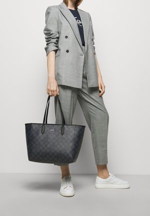 Donna che indossa un completo grigio e sneakers bianche, mentre tiene una grande borsa a rete nera con manici doppi.