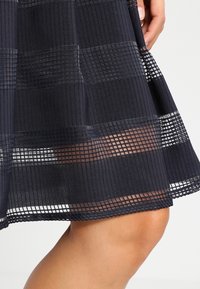 Svart lagerklänning med ett strukturerat rutmönster, som har genomskinliga och ogenomskinliga sektioner, samt en utsvängd nederdel.