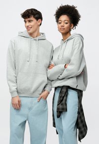 Lysegrå hettegensere med kengurulommer, utstyrt med snorer og ribbede mansjetter, brukt sammen med lyseblå jeans og en svart rutete skjorte knyttet rundt livet.