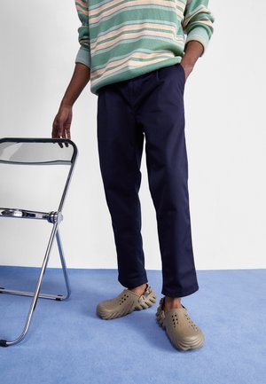 Personne portant un pantalon bleu marine, des chaussures beiges à enfiler et un pull rayé vert, debout à côté d'une chaise pliante en métal sur un tapis bleu.