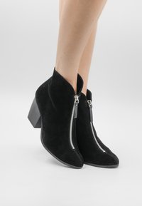 Botine negre din suede până la gleznă, cu vârf ascu�țit, fermoare laterale și toc gros. Textura este moale, iar designul prezintă linii clare.