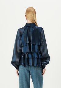Blouse en satin marine avec des volants superposés, col haut et manches bouffantes longues, associée à un jean bleu clair, vue de dos.