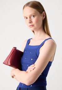 Haut texturé bleu avec des embellissements décoratifs, associé à un petit sac en cuir rouge. Le haut présente des bretelles larges et une coupe ajustée.