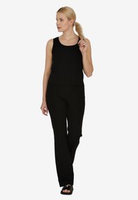 Kobieta z blond końskim ogonem, ubrana w czarną bezrękawnik, dopasowane czarne spodnie i czarne sandały, idzie i patrzy w lewo.