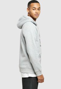 Hanorac gri cu glugă, dotat cu un buzunar tip "kangaroo", manșete și tiv cu model în relief. Este purtat peste un tricou alb și are un design simplu, casual.
