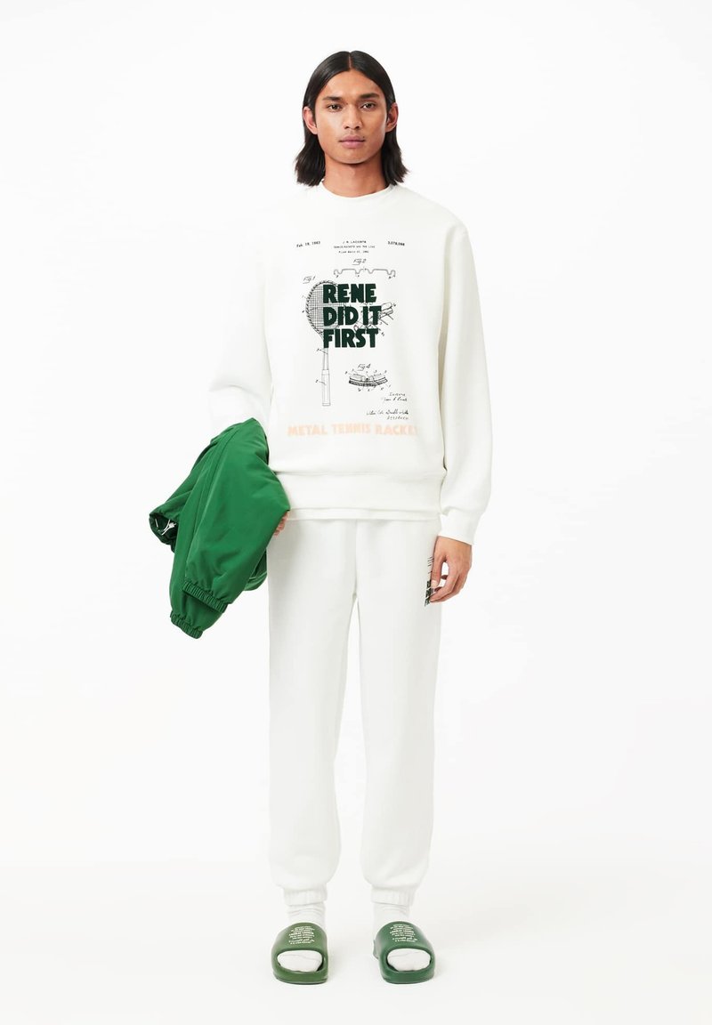 Un sweat-shirt blanc avec des graphismes verts, un pantalon de survêtement blanc assorti et une veste bomber verte, le tout complété par des sandales vertes à enfiler.