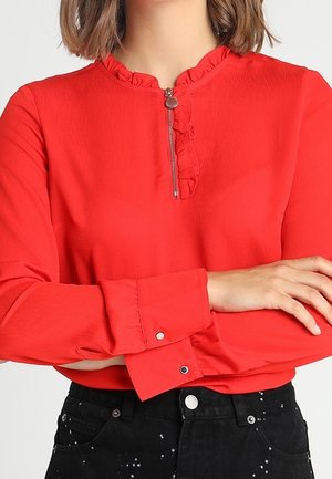 Blusa roja de manga larga con cuello fruncido y cremallera, usada metida dentro de pantalones negros de tiro alto con detalles de salpicaduras blancas de pintura.