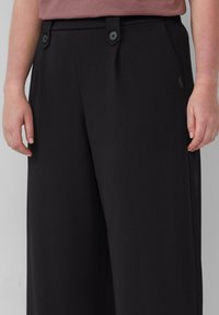 Schwarze Hose aus glattem Stoff, mit flachem Vorderteil, zwei Knöpfen an der Taille und seitlichen Taschen. Taillierter, weiter Schnitt.