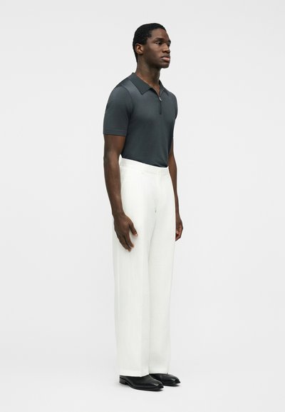 Homme debout portant un polo gris foncé à manches courtes et un pantalon taille haute blanc avec des chaussures noires, sur un fond clair uni.