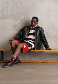 Μαύρο δερμάτινο μπουφάν πάνω από ένα μαρτούν και άσπρο ριγέ πουκάμισο, κόκκινες βερμούδες με μαύρες λεπτομέρειες, μαύρα παπούτσια, καθισμένος σε μια ξύλινη παγκάκι.