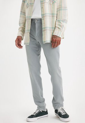 Mann trägt hellgraue Hose, schwarze Turnschuhe mit weißen Sohlen und Schnürsenkeln sowie ein ungeknotetes kariertes Hemd über einem weißen Oberteil.