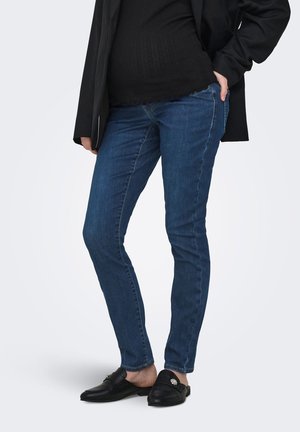 Person iført tætsiddende blå jeans, sort top, sort blazer og sorte slip-on loafers, stående med den ene hånd i lommen.