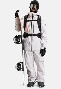 Person in weißer Snowboardausrüstung, schwarzen Handschuhen und einer Skibrille, der ein Snowboard aufrecht mit Bindungen vor einem weißen Hintergrund hält.