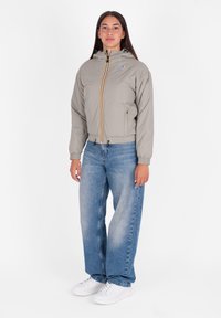 Giacca corta grigia chiara con cappuccio, dotata di zip frontale e strisce colorate. Indossata con jeans larghi blu e sneaker bianche.