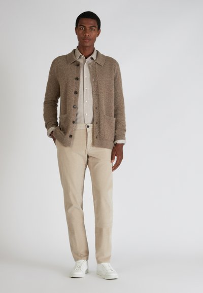 Beige gebreide cardigan met vier knopen, twee voorzakken; lichtgrijs overhemd met knopen eronder; crèmekleurige gestreepte broek; witte sneakers.