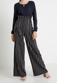 Námořnicky modrý top s dlouhými rukávy a průstřihem, doplněný o kalhoty s vysokým pasem a širokými nohavicemi s vertikálními bílými pruhy.