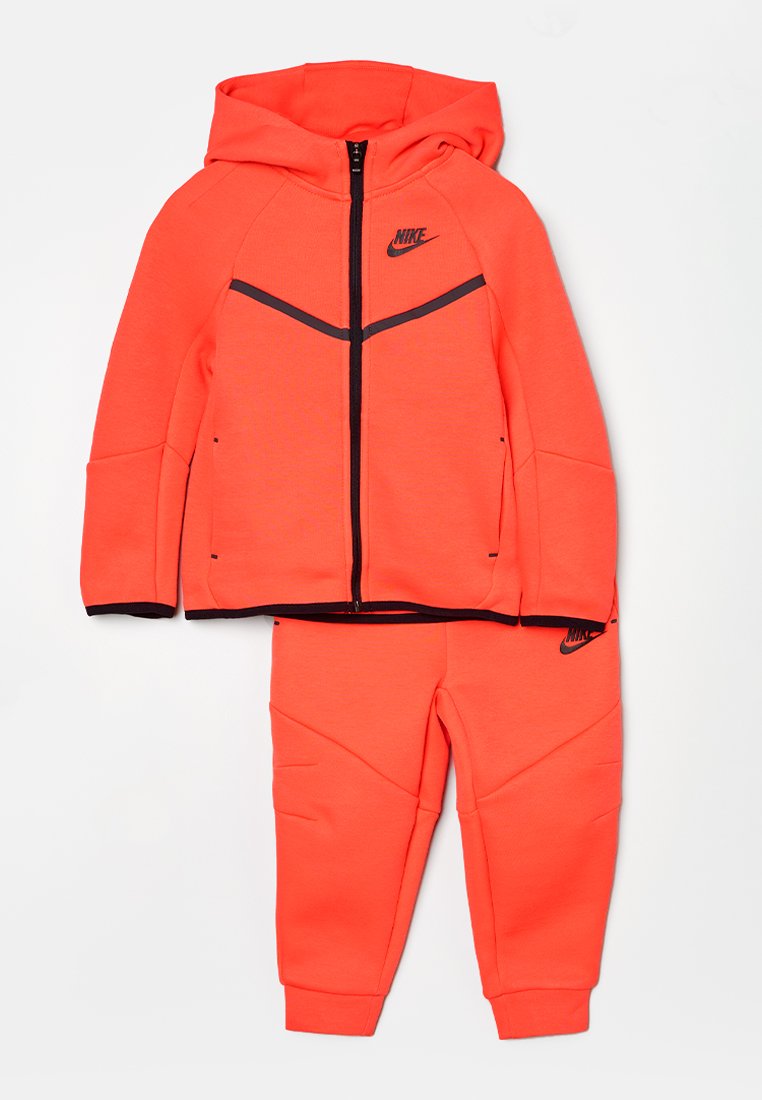 Dres Nike w jasnym neonowym pomarańczowym kolorze, składający się z zamykanej na zamek bluzy z kapturem z czarnymi akcentami oraz dopasowanych spodni z teksturowanym wykończeniem i mankietami.