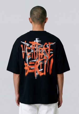 Persoon met geschoren hoofd die een zwart oversized T-shirt draagt met rode en witte graffiti-achtige tekst en ontwerp op de achterkant.