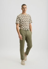 Camisa de manga corta beige con un patrón de zigzag marrón, combinada con pantalones verdes oliva y zapatos beige claros; un conjunto con cinturón y un corte relajado.