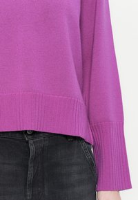 Pull en tricot violet avec des détails de bord côtes à l'ourlet et aux poignets, au fit décontracté, porté avec un jean gris foncé.