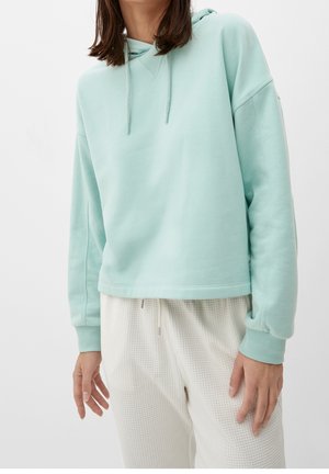 Personne portant un sweat à capuche bleu clair avec cordons et un pantalon blanc texturé, debout les bras détendus devant un fond uni.
