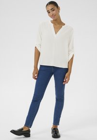 Blusa bianca a maniche corte con scollatura a V e risvolti, abbinata a jeans skinny blu scuro e mocassini neri. Texture liscia, vestibilità casual.